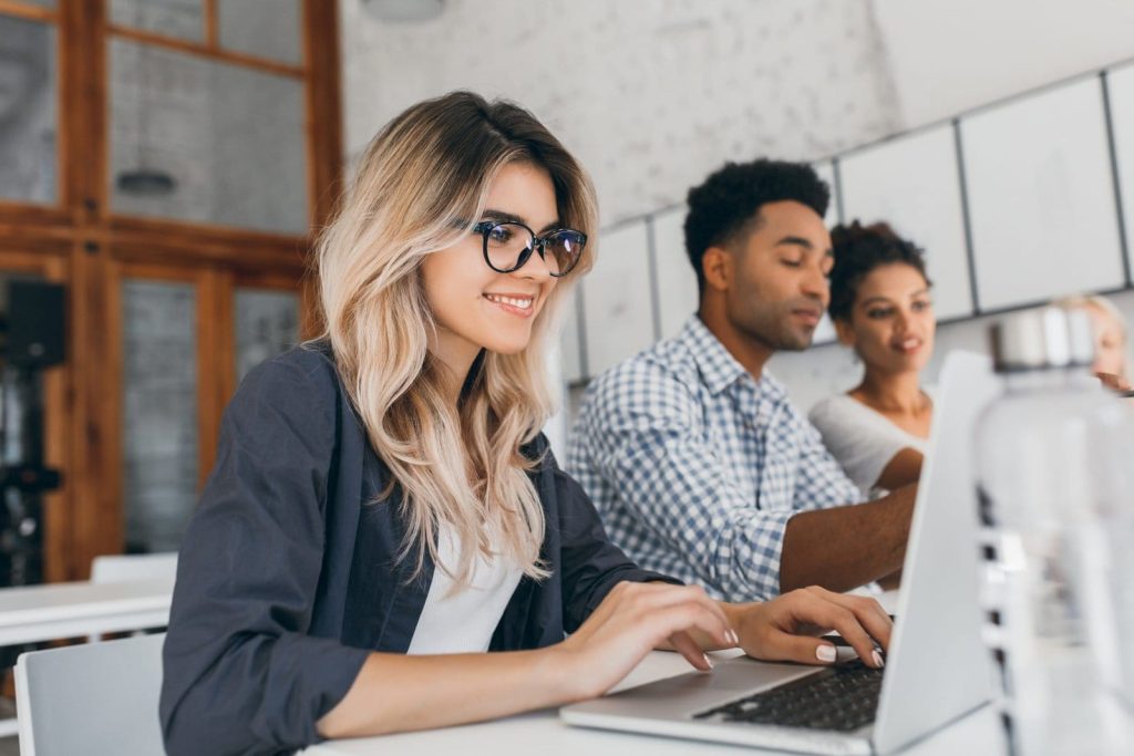 beautiful curly female freelancer with cute manicure using laptop smiling indoor portrait blonde secretary sitting beside african coworker blue shirt scaled 1