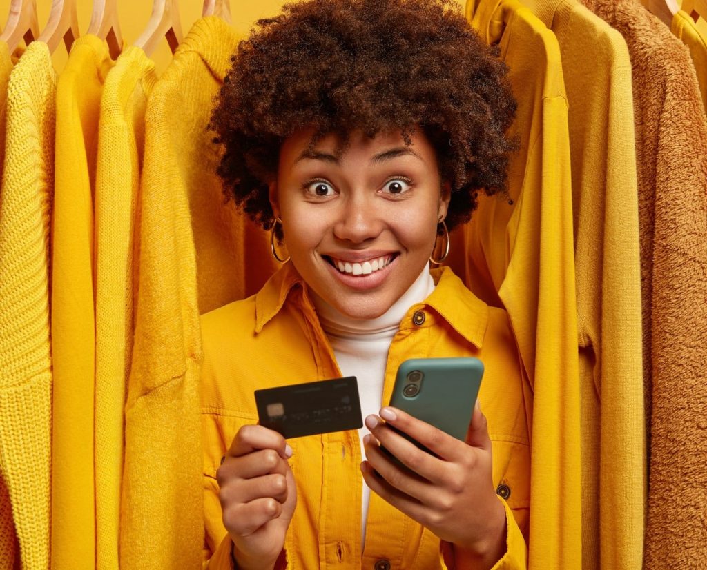 positive mixed race shopaholic stands near rack with clothes uses credit card smartphone making purchasing